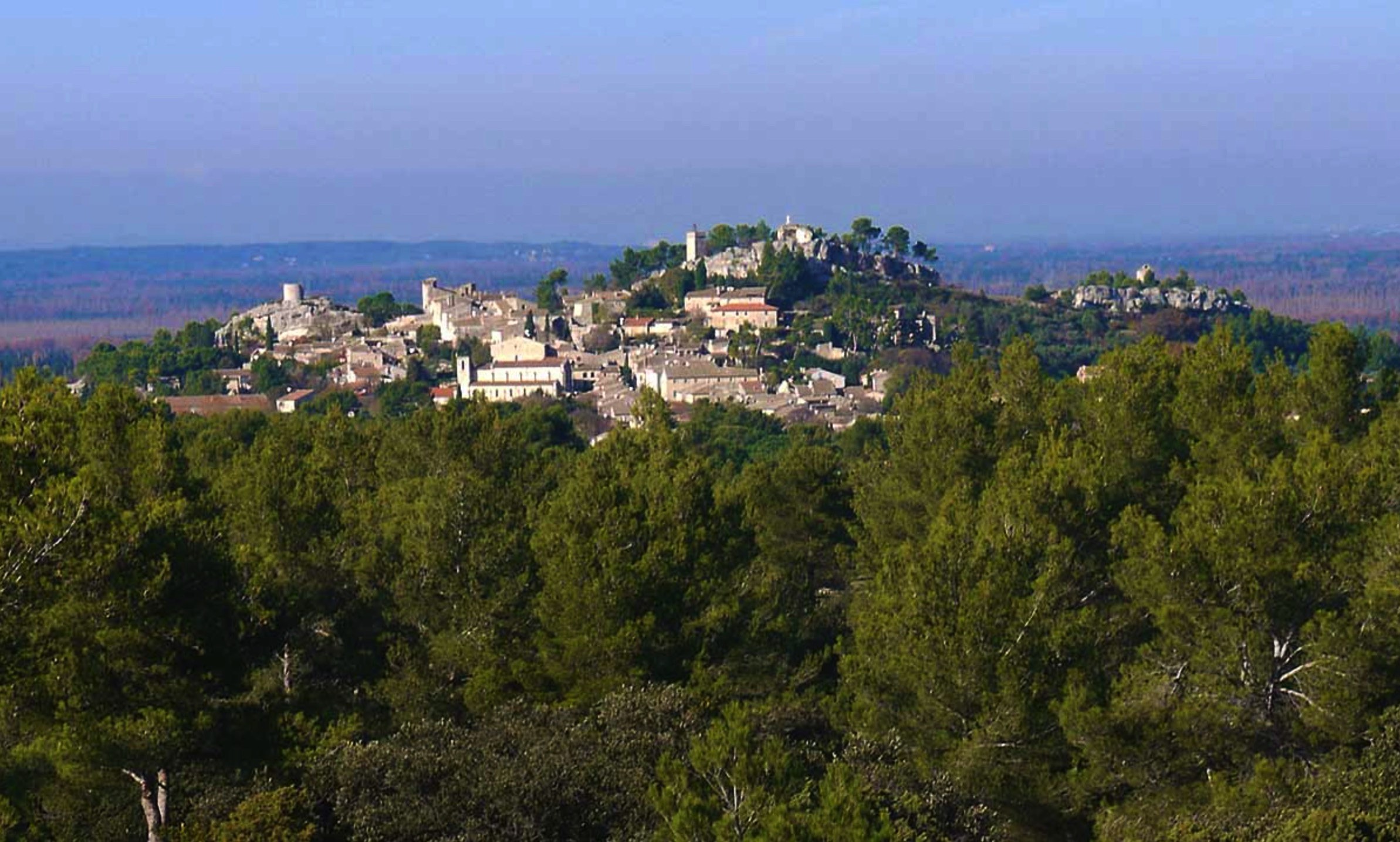 A scenic view of a historic village perched on a hilltop surrounded by lush green forests. The village features tightly clustered stone buildings.